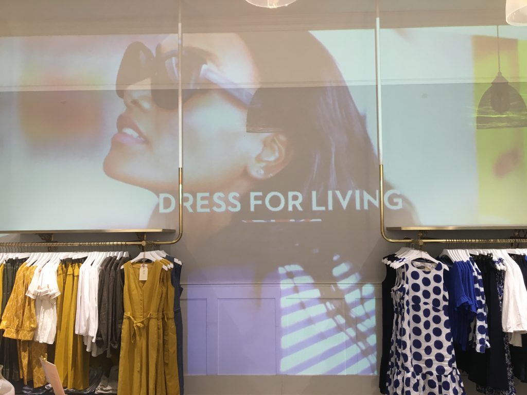 Photo of creative projection displayed on the back wall of a fashionable clothing store. The projection is a profile shot of a woman’s face wearing sunglasses with the caption “dress for living” displayed over the top