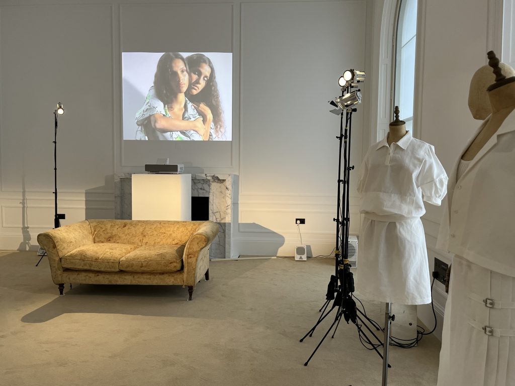 Photo of creative projection containing two women hugging displayed on a white wall. The room has a cream sofa, some lighting, and mannequins displaying fashionable women’s clothing
