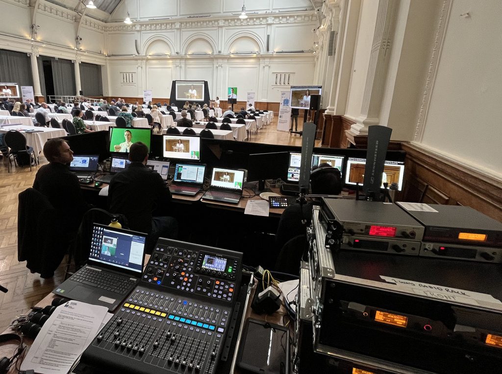 Photo of a conference room with an audio desk and wireless equipment in the foreground, and a conference happening in the background. The conference shows multiple people using laptops and facing towards a head table with