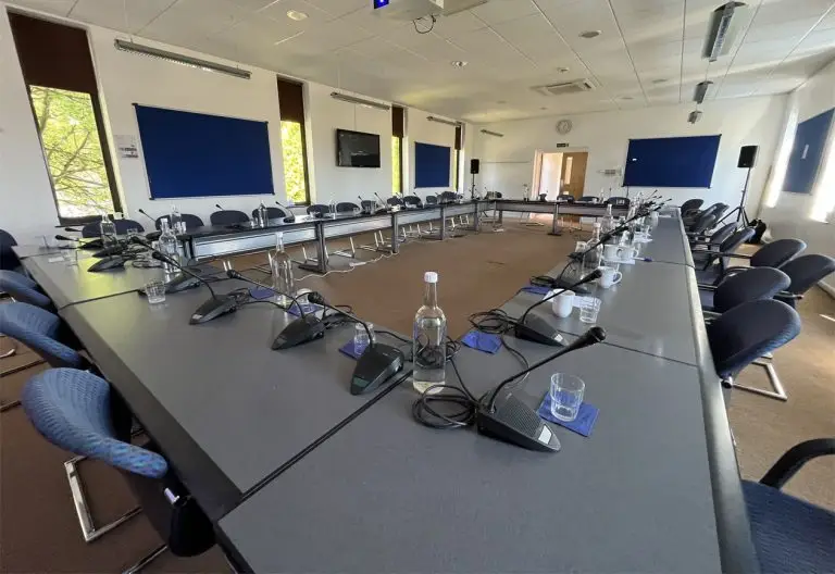 Photo of long tables arranged in a rectangle in a conference room. A push-to-talk microphone is positioned in front of each chair